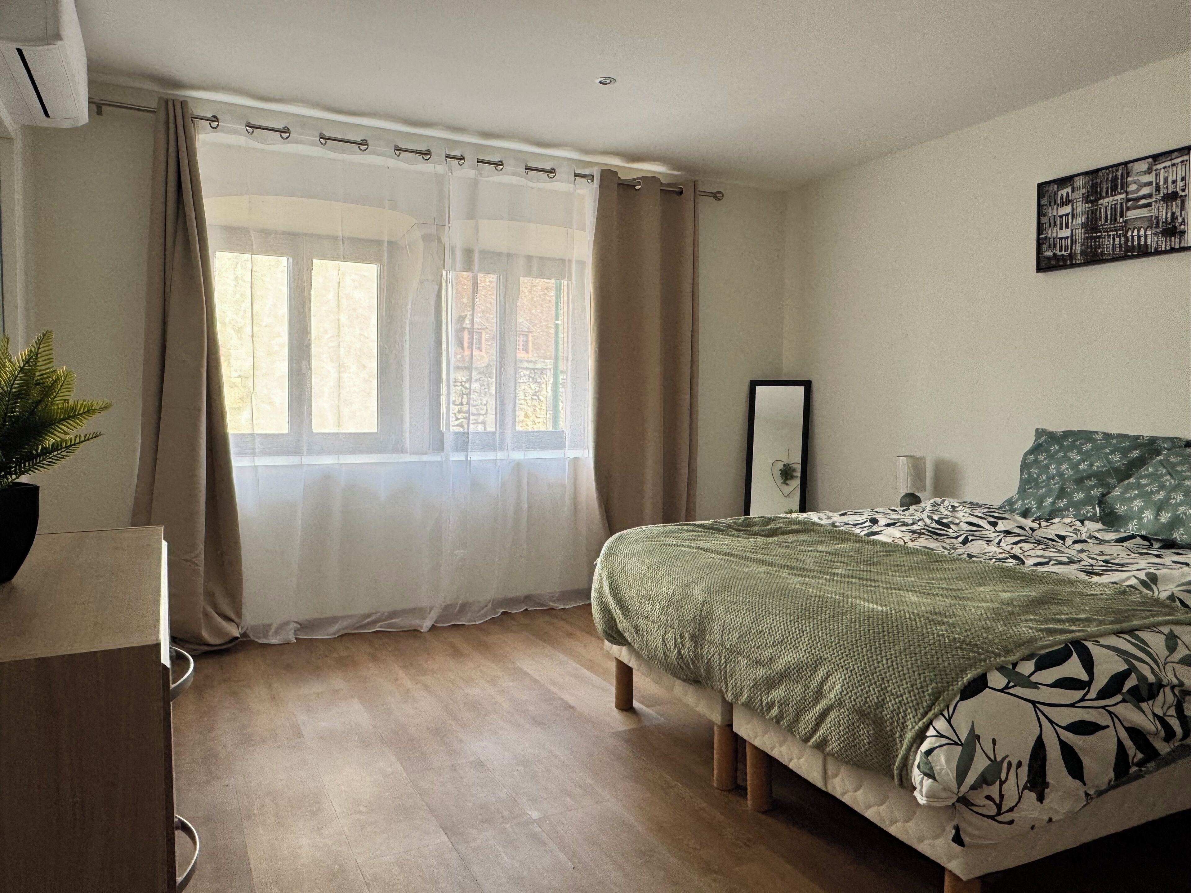 A bedroom with a bed, a window with curtains, a plant, and a wall-mounted mirror.