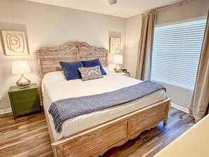 A bedroom with a wooden bed, two nightstands, a lamp, a clock, and a window with blinds. - The Scenic Retreat at Ciboney 4010 - Gulfview - 3-bedroom - Pet Friendly (Miramar Beach)