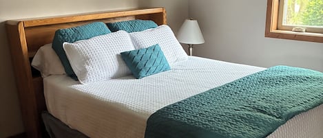 Una cama bien hecha con cabecera de madera, una lámpara de noche y una ventana con persianas.