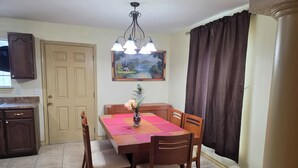 A dining area with a wooden table, chairs, a vase with flowers, a painting on the wall, and a ceiling light fixture.