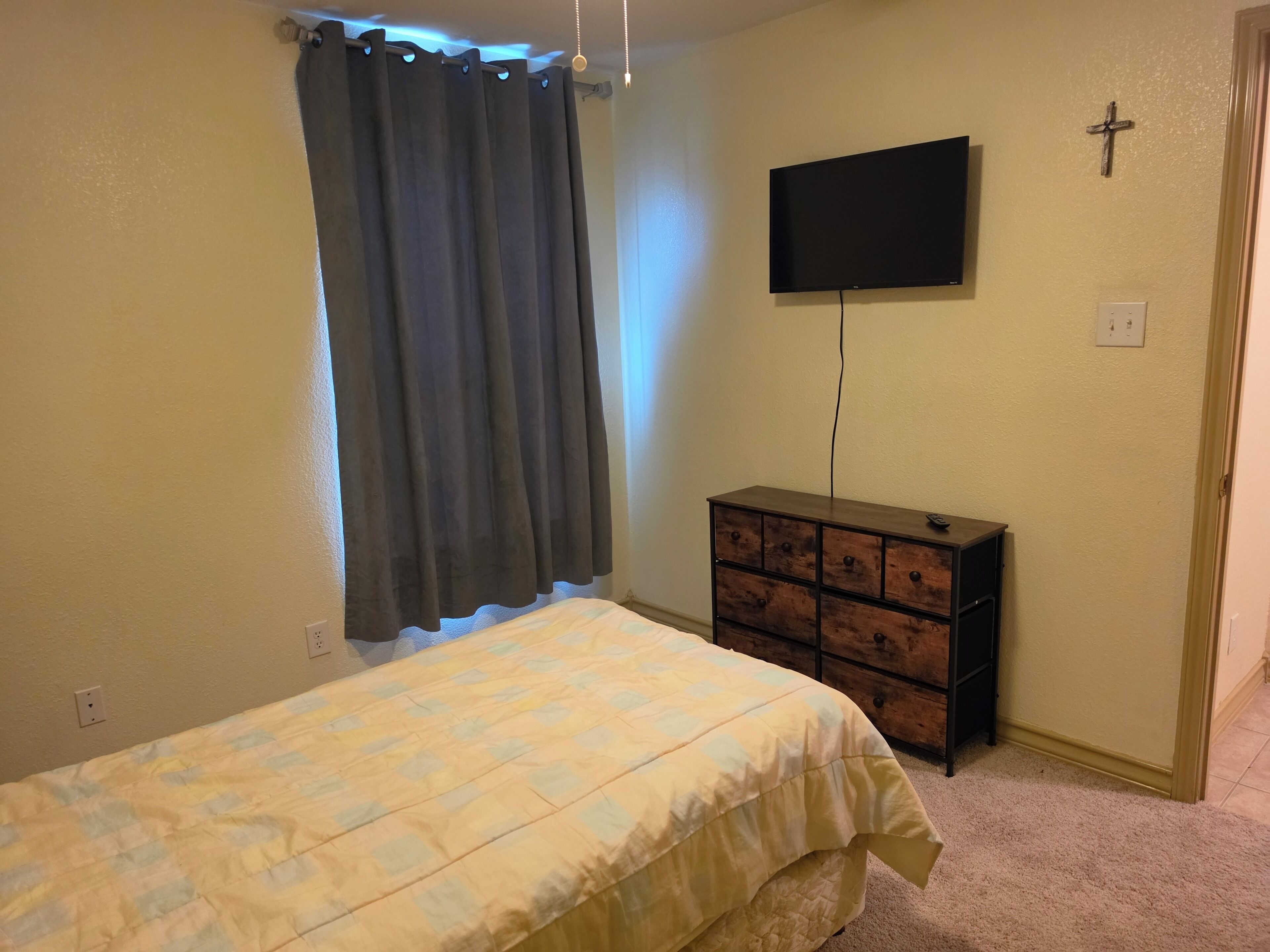 Un dormitorio con una cama, un aparador, un televisor en la pared y una cruz decorativa.