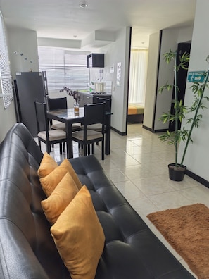 A modern living room with a black sofa, a dining table, and chairs. There is a kitchen area in the background with a refrigerator, stove, and microwave. A potted plant is visible on the floor. - Apartahotel a 3 minutos del Parque del Cafe (Pereira)