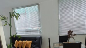 Ein Zimmer mit einer schwarzen Couch, einem Esstisch mit Stühlen und zwei Fenstern mit Jalousien.
