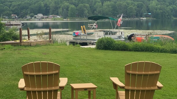 Two wooden Adirondack chairs facing a lake with boats, a dock, and a forested hill in the background. - Pet friendly Lakefront House with hot tub and large Deck on Summit Lake (Argyle)