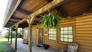 A wooden cabin with a porch, a welcome sign, a rocking chair, and a hanging fern.