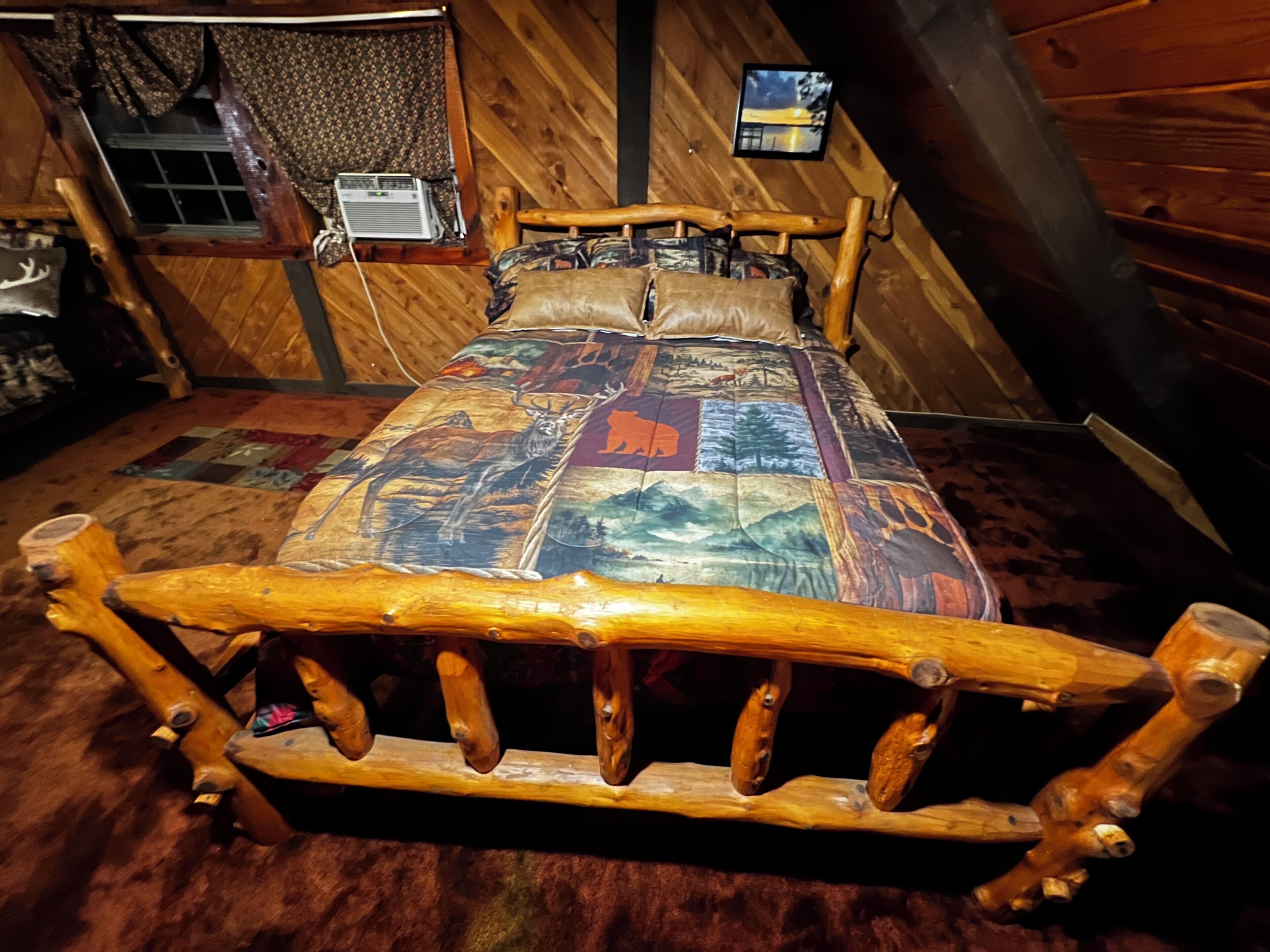Een houten bed met hoofdeinde, een sprei met een boslandschap, een raam met gordijnen en een aan de muur hangend schilderij.