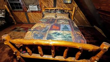Een houten bed met hoofdeinde, een sprei met een boslandschap, een raam met gordijnen en een aan de muur hangend schilderij.