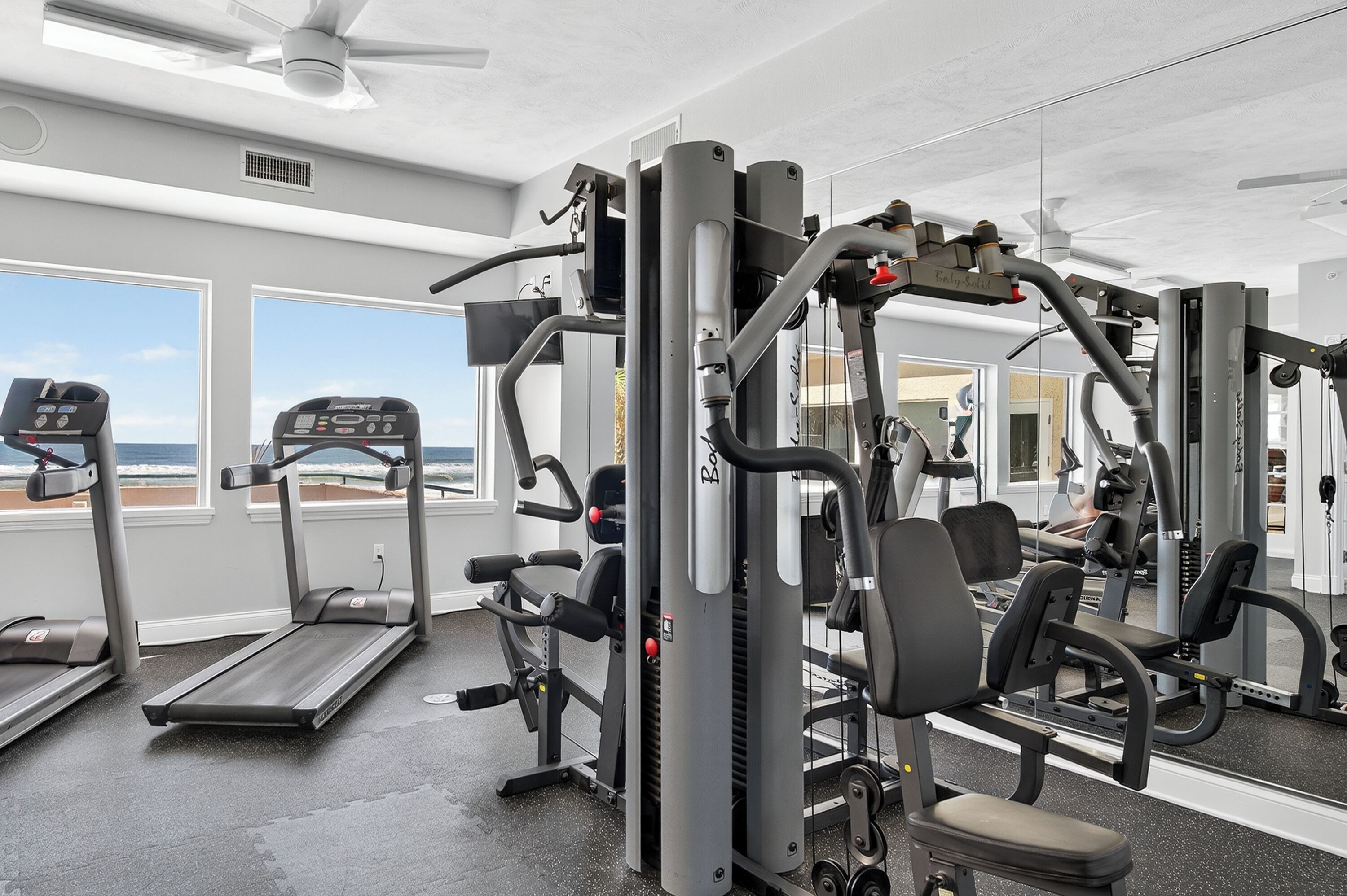 Une salle de sport équipée de tapis de course, d’appareils de musculation et dotée d’une grande fenêtre donnant sur la plage.