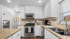 Una cocina moderna con armarios blancos, encimeras de granito, electrodomésticos de acero inoxidable y un amplio ventanal con persianas.