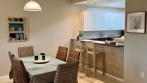 Een eetgedeelte met rieten stoelen, een tafel met tafelonderzetter, een hanglamp en een keuken op de achtergrond.