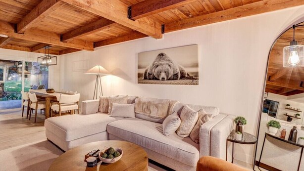 A modern living room with a wooden ceiling, a sofa, a coffee table, and a dining area with a lamp.