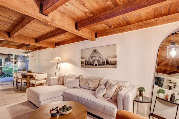 A modern living room with a wooden ceiling, a sofa, a coffee table, and a dining area with a lamp.