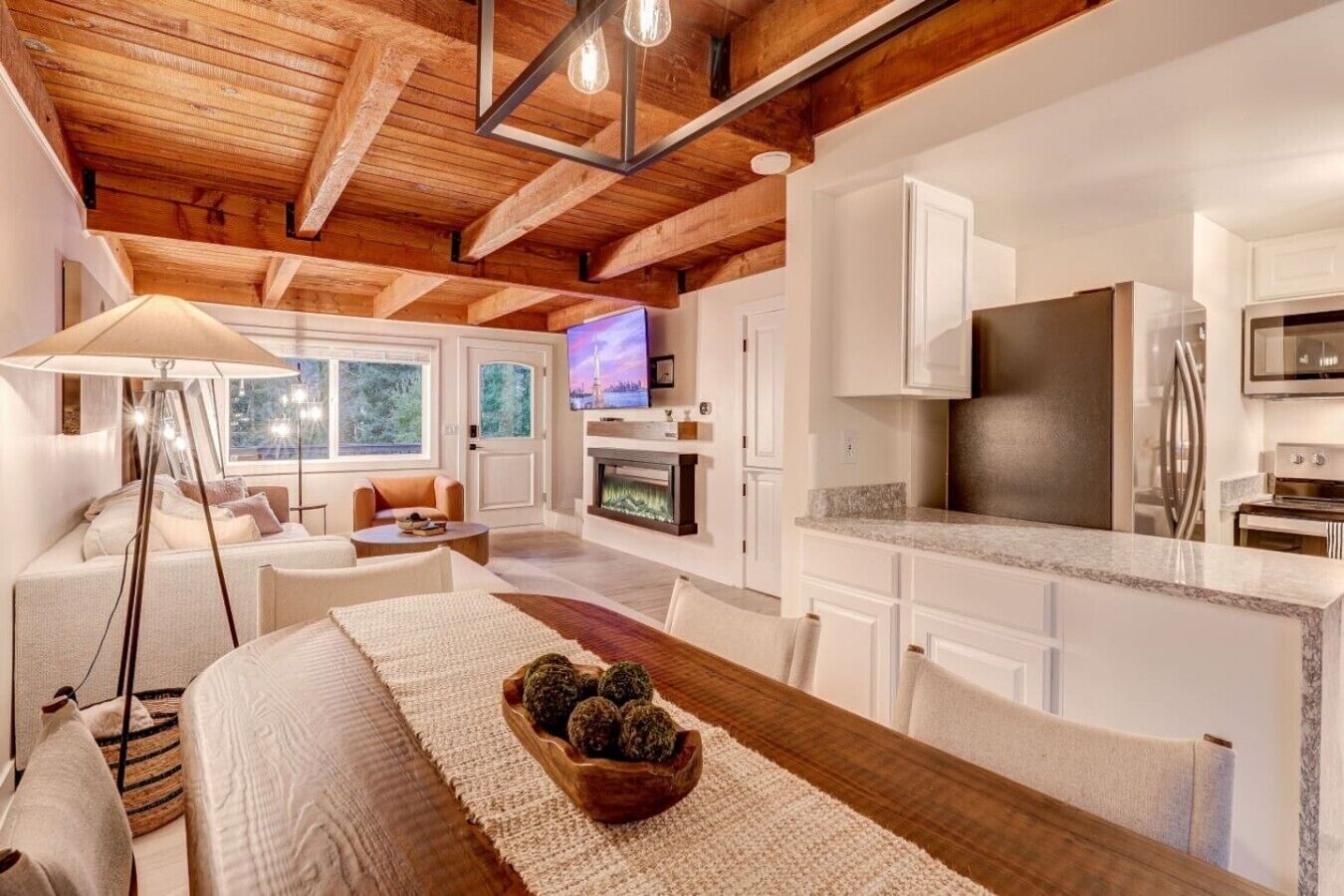 Ein modernes Interieur mit Holzbalkendecke, einem Essbereich mit Tisch und Stühlen, einer Küche mit weißen Schränken und einem Fernseher im Hintergrund.