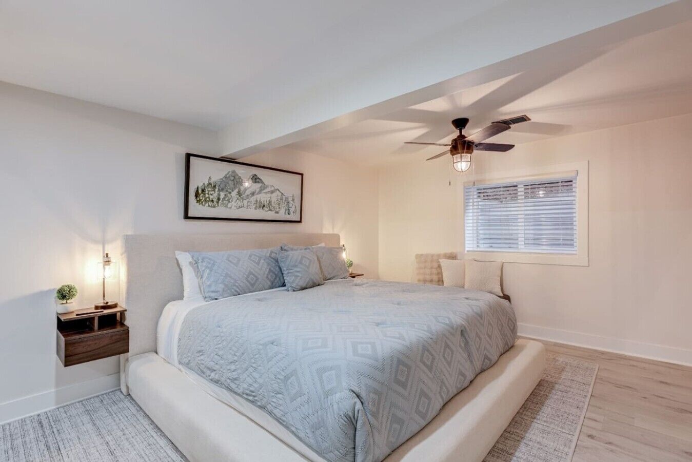 Une chambre à coucher avec un lit, un ventilateur de plafond, une fenêtre avec des stores, une table de chevet avec une lampe et un tableau encadré au mur.