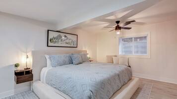 Une chambre à coucher avec un lit, un ventilateur de plafond, une fenêtre avec des stores, une table de chevet avec une lampe et un tableau encadré au mur.
