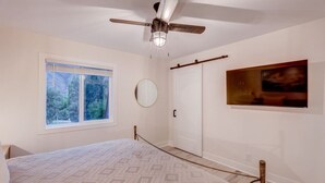 A bedroom with a bed, a ceiling fan, a window with a view, a door, and a painting on the wall.