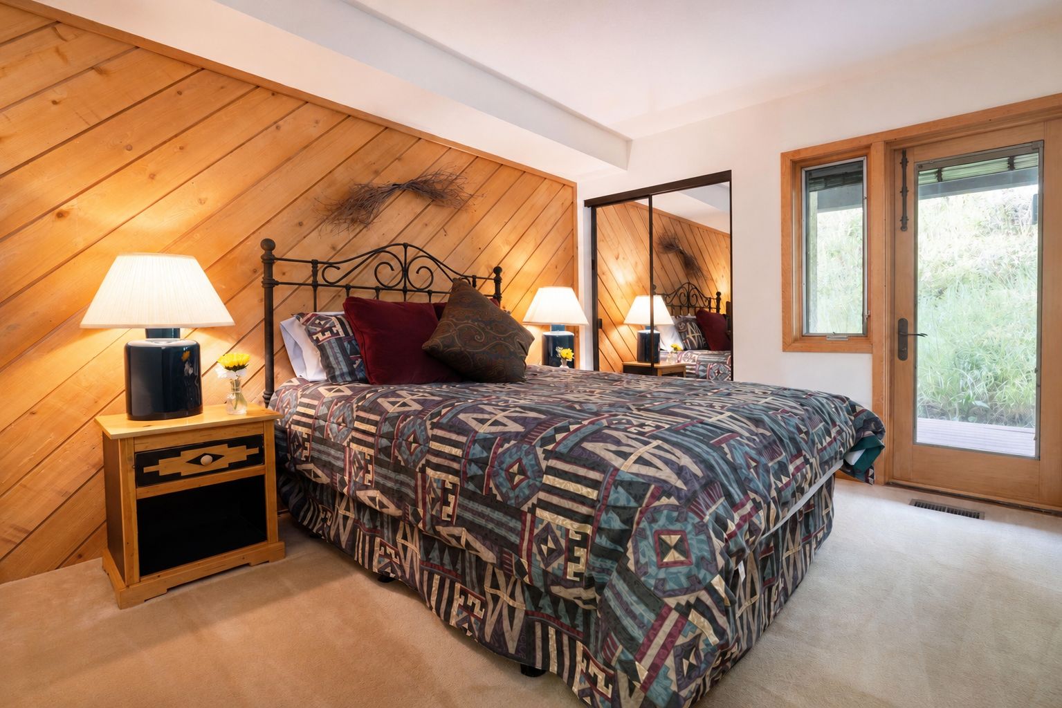Ein Schlafzimmer mit Holzbalkendecke, einem Bett mit gemusterten Betttextilien, zwei Nachttischlampen, einem Nachttisch mit Schublade und einem großen Fenster mit Blick auf Grünflächen.