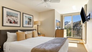 Habitación de hotel con una cama grande, una mesita de noche, una lámpara, un televisor montado en la pared y un balcón con vistas a la montaña.