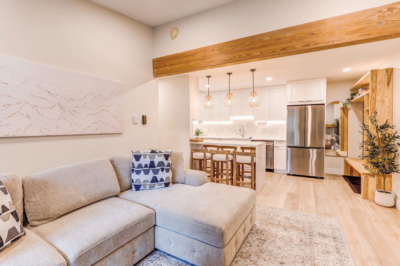 Una sala moderna con un sofá modular, un comedor con sillas de madera, una cocina con armarios blancos y electrodomésticos de acero inoxidable, y una viga de techo de madera.