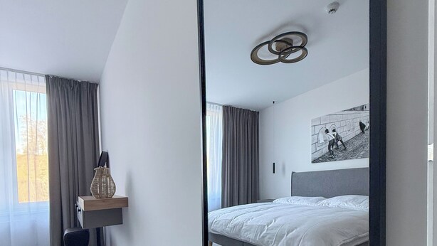 Een moderne slaapkamer met een grote spiegel, een bed, een bureau met stoel en een klein krukje.