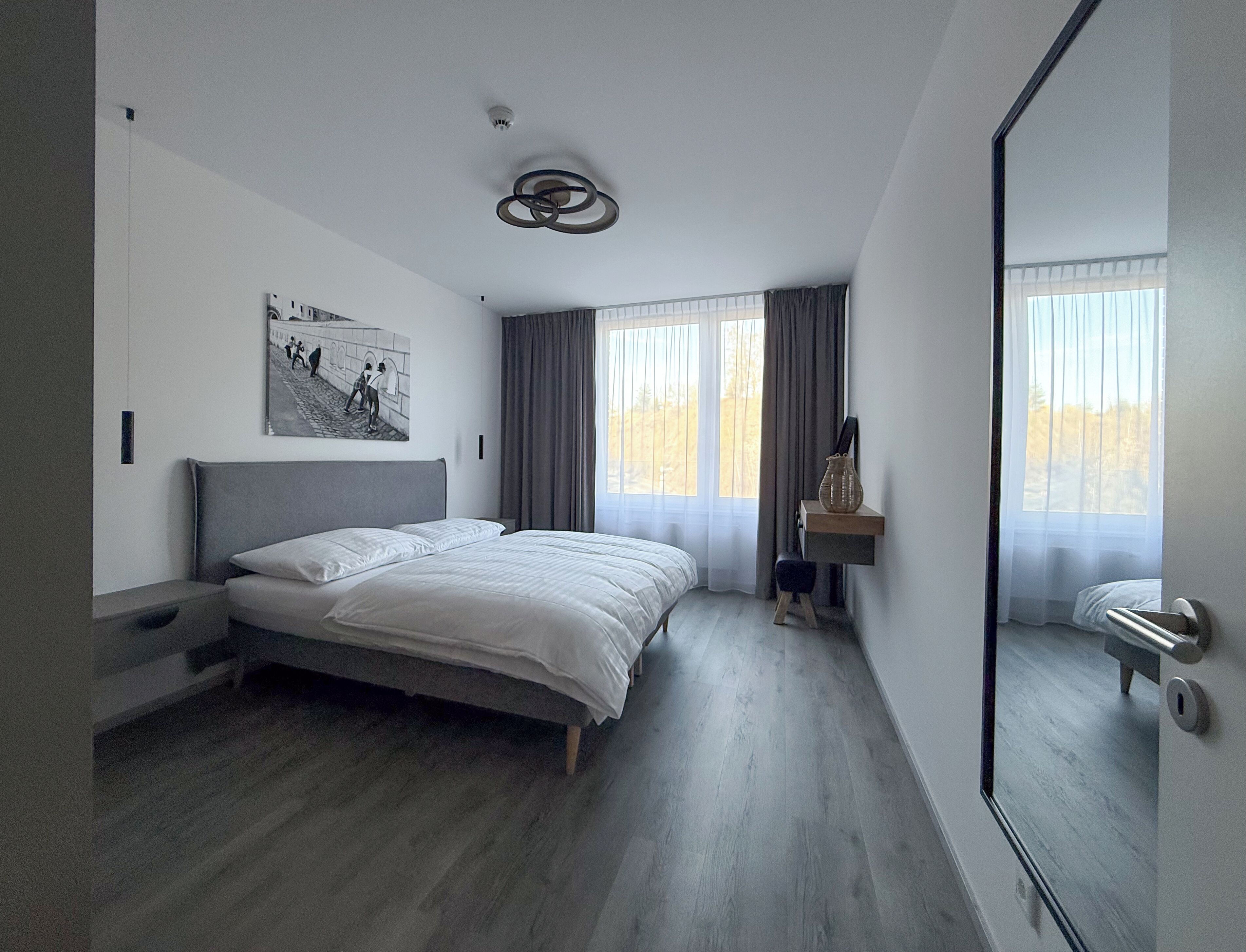 Une chambre à coucher moderne avec un lit, un tableau, un bureau agrémenté d’un vase et un grand miroir.