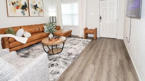 A living room with a brown leather sofa, a round wooden coffee table, a white door, and a ceiling fan. - Haven on the Hill, Modern 3BR Near Fort Cavazos (Copperas Cove)