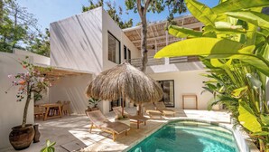 Un espace piscine extérieur moderne, doté de chaises longues, d’un parasol en chaume et de plantes en pot.
