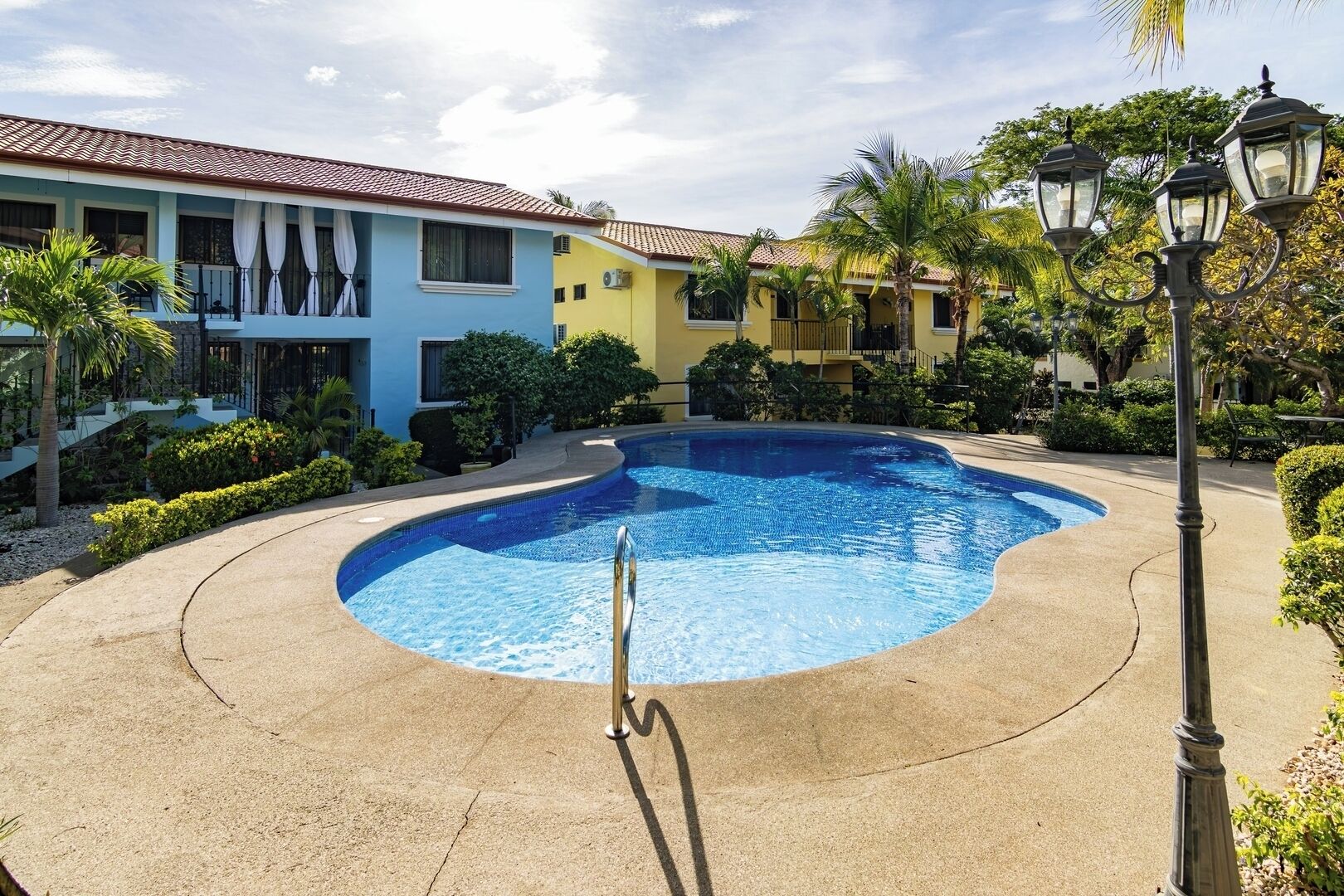 Una piscina de borde curvo, rodeada por una terraza de concreto, junto a un edificio con balcones y techo de tejas.