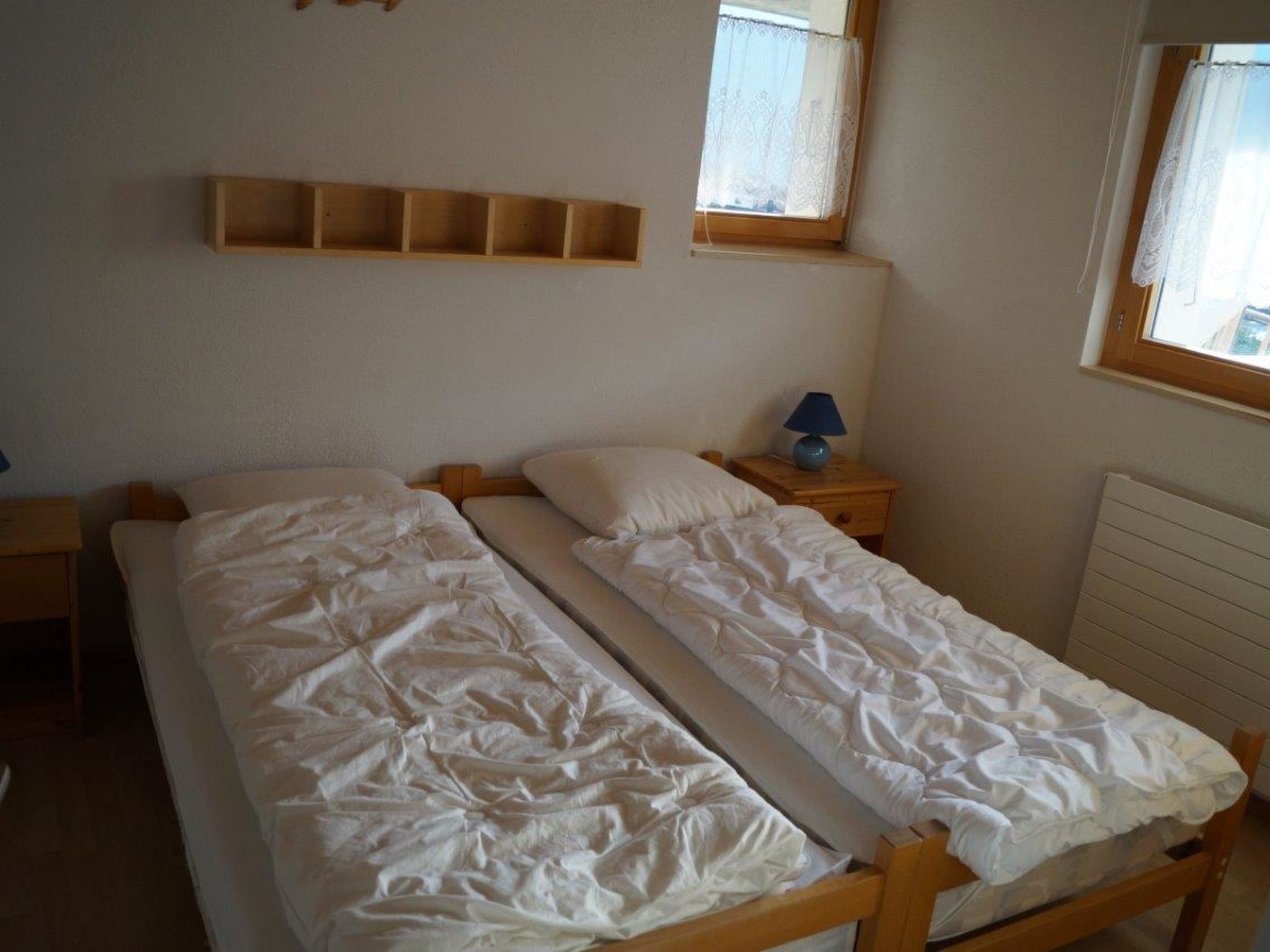 Una habitación con dos camas individuales, una mesita de noche de madera con lámpara, un estante sobre las camas y una ventana con cortina de encaje.
