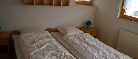 Una habitación con dos camas individuales, una mesita de noche de madera con lámpara, un estante sobre las camas y una ventana con cortina de encaje.