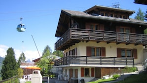 A two-story building with a balcony, a ski lift in the background, and a grassy area with dandelions. - Dilaila, (Grächen), by Interhome (Grächen)