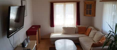 Uma sala de estar acolhedora com televisão de tela plana, um sofá, uma mesinha e uma janela com cortinas.