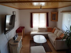 Ein Wohnzimmer mit einer Couch, einem Fernseher, einem kleinen Tisch und einem Fenster mit Vorhängen.