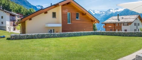 Una cabaña de madera con base de piedra, rodeada de un césped cuidado y con montañas al fondo.