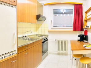 Eine kompakte Küche mit Herd, Spüle und Backofen. Ein Fenster mit rotem Vorhang, ein Heizkörper und ein kleiner Tisch mit Stühlen sind ebenfalls vorhanden.