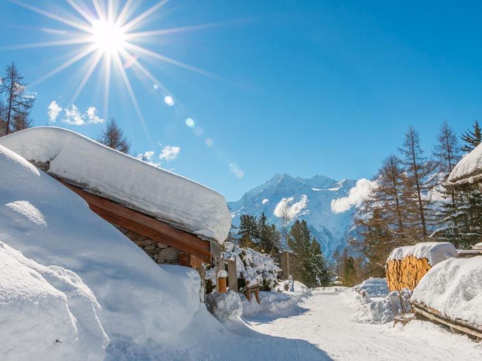 Ein verschneiter Bergpfad führt zu einer Holzhütte mit Steinunterbau, umgeben von schneebedeckten Bäumen und Bergen im Hintergrund.