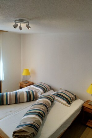 A single bed with striped bedding, a bedside table with a lamp, and a ceiling light fixture. - Nr. 20 - Haus Opal, (Grächen), by Interhome (Grächen)