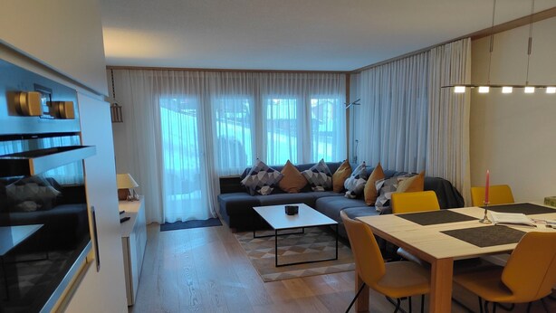 Una sala de estar moderna con sofá, mesa de comedor y sillas, una zona de cocina y ventanas grandes con cortinas.