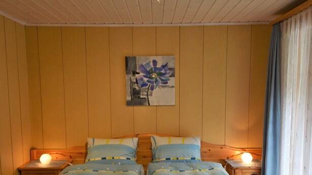 Ein Doppelzimmer mit Holzwandverkleidung, zwei Betten mit passendem blau-gelben Bettzeug, einem Bild an der Wand und einem Nachttisch mit Lampe auf jeder Seite.