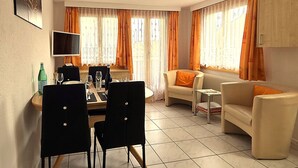 Ein möbliertes Wohnzimmer mit Essbereich, großen Fenstern inklusive Vorhängen, einem Flachbildfernseher und einer Heizung.