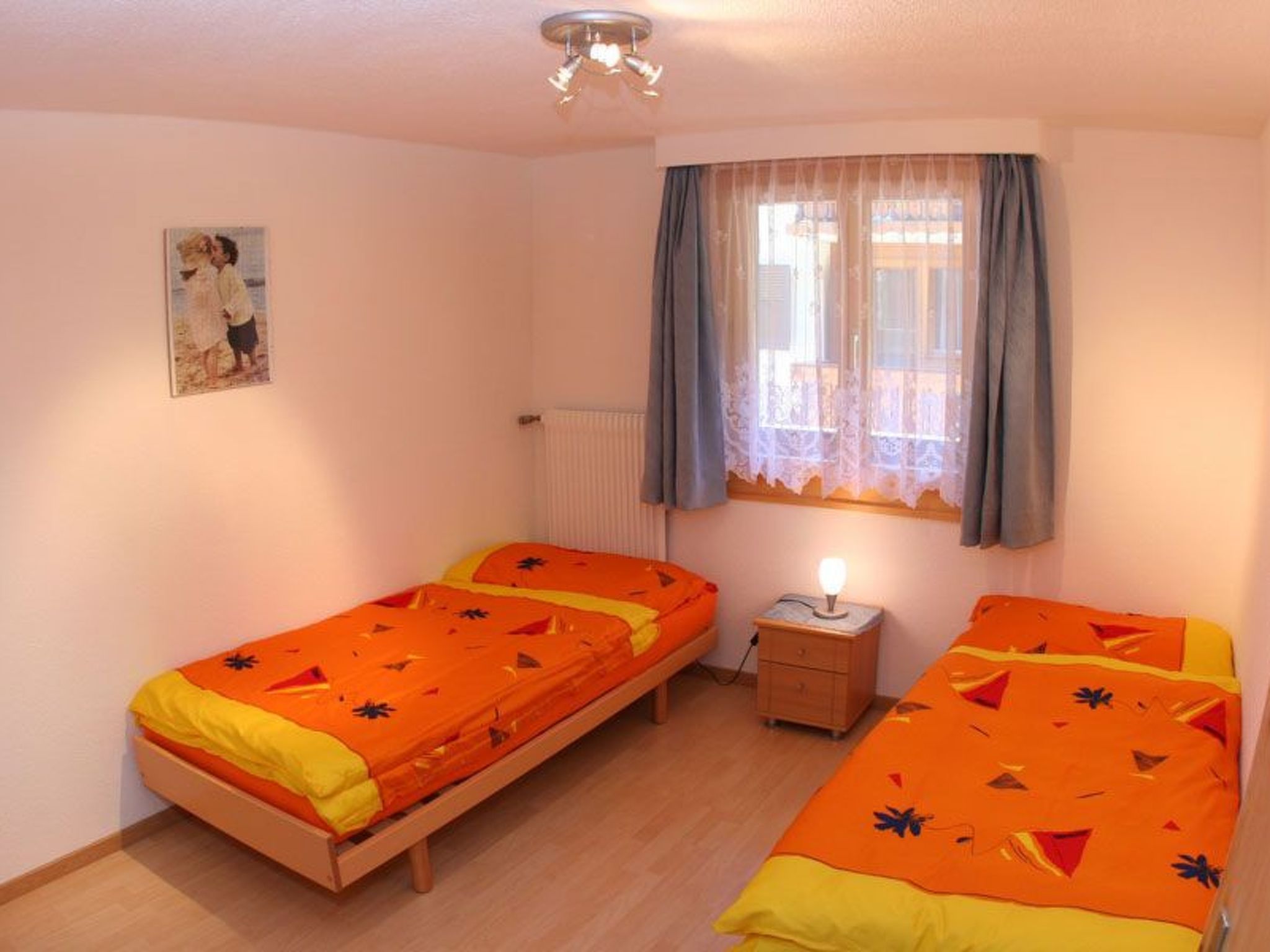 Una habitación con dos camas individuales, una mesita de noche, una lámpara, un radiador y una ventana con cortinas.