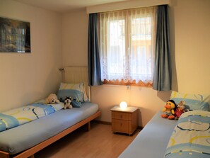 A bedroom with two beds, a nightstand with a lamp, and a window with curtains. - Ferienwohnungen Zur Heimat, (Grächen), by Interhome (Gr ächen)