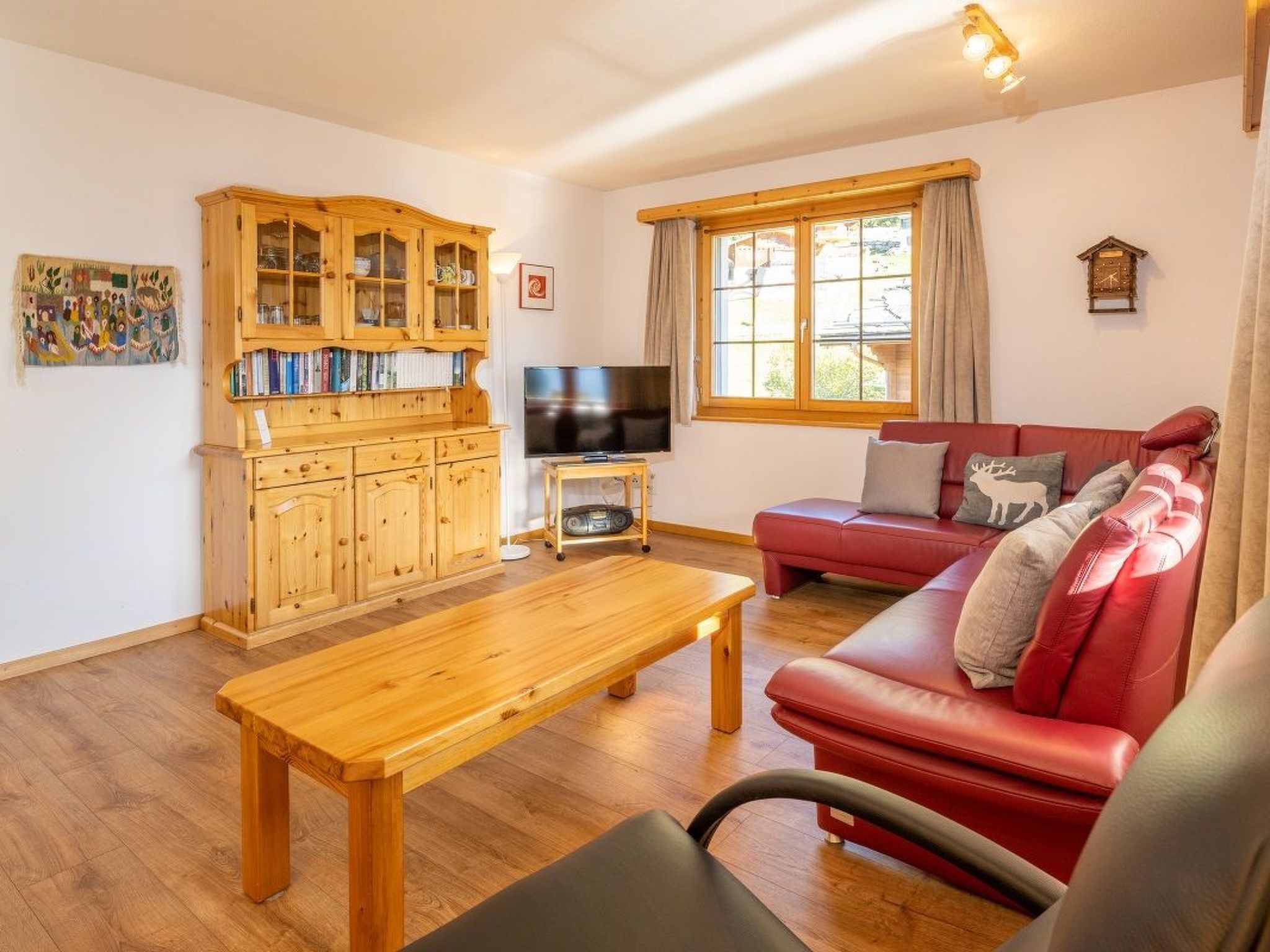 A cozy living room with a wooden coffee table, a red sofa, a television, and a wooden cabinet with glass doors.