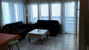 Ein Wohnzimmer mit einer Couch, einem Tisch und einem Sessel. Es gibt Fenster mit Vorhängen.