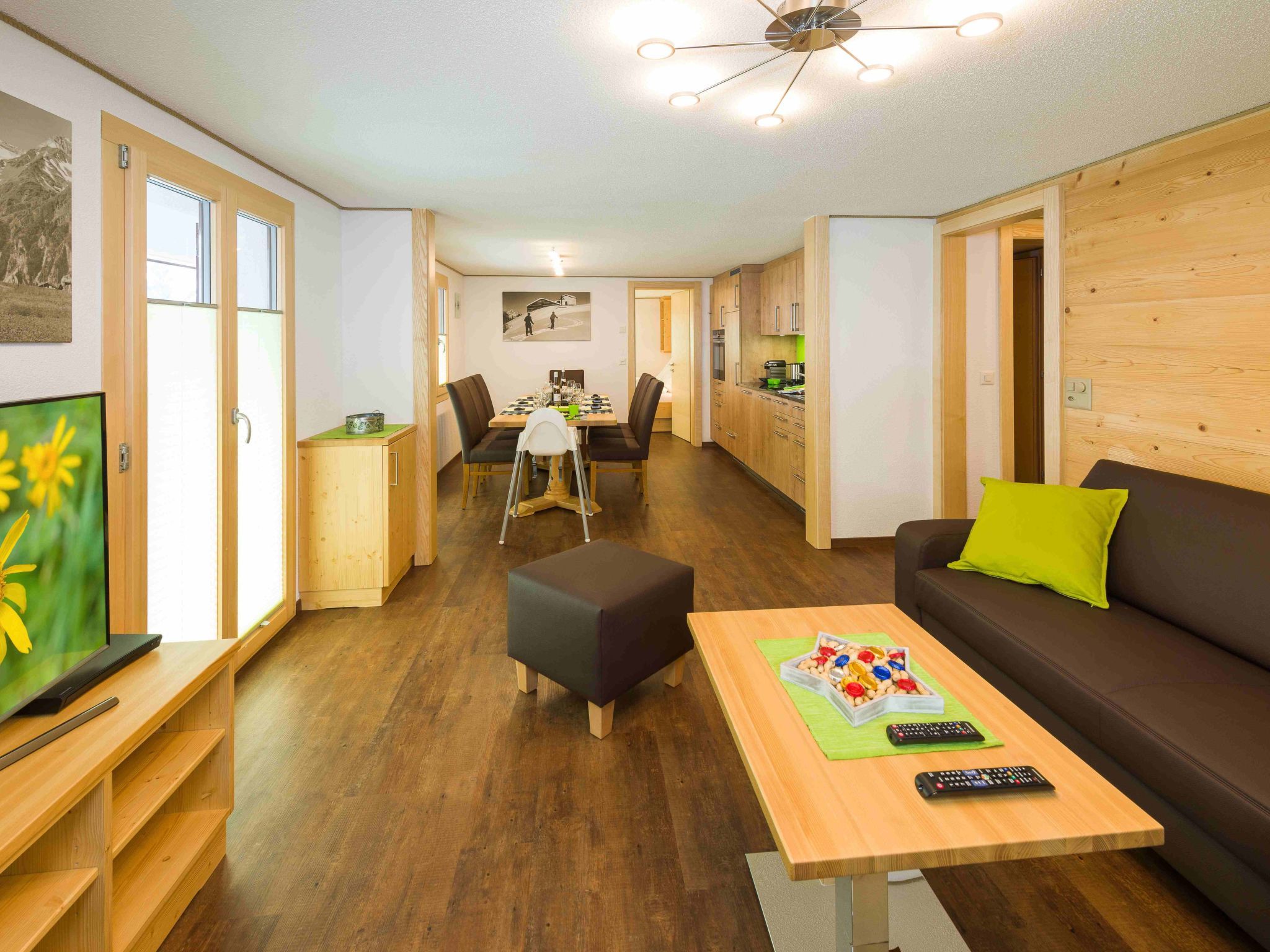 A modern living room with a sofa, a coffee table, and a TV. There is a dining area with a table and chairs. The room has wooden flooring and a kitchen area with cabinets.