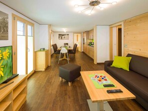 A modern living room with a sofa, a coffee table, and a TV. There is a dining area with a table and chairs. The room has wooden flooring and a kitchen area with cabinets. - Ferienwohnungen Lunic, AZapartments.ch, B&B, (Grächen), 1057 by Interhome (Grächen)