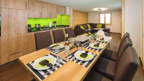 Eine moderne Küche mit Holzausstattung, eine Essecke mit einem langen Tisch für sechs Personen sowie eine Sitzecke mit einem braunen Sofa und einem kleinen Tisch.