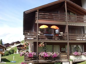 Ein mehrstöckiges Holzgebäude mit Balkonen, Blumenarrangements im Freien und blauem Himmel.