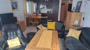 Een woonkamer met een houten salontafel, een zwarte leren bank en een eethoek, met een keuken op de achtergrond.