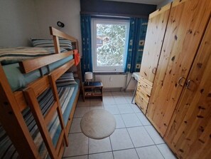 Ein kleines Zimmer mit Etagenbetten, einem Tisch, einer Lampe und einem Kleiderschrank.